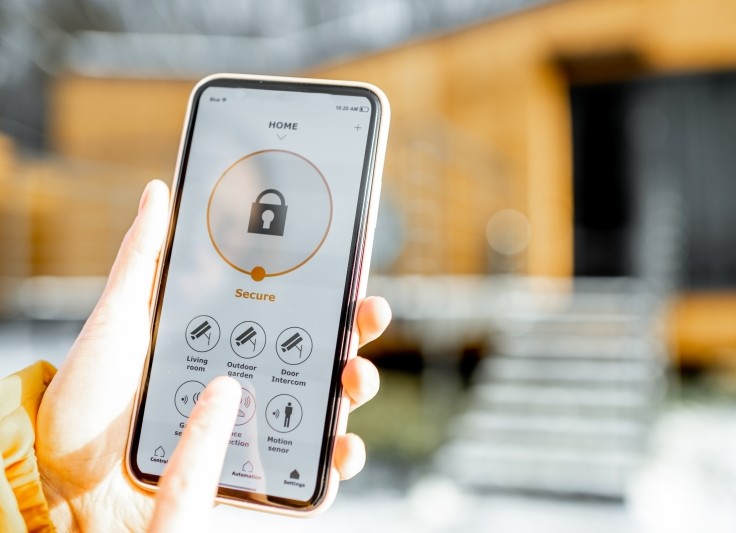A person holds a smartphone displaying a home security app with the system set to “Secure,” showing controls for cameras, motion sensors, and other smart home features, with a house blurred in the background.