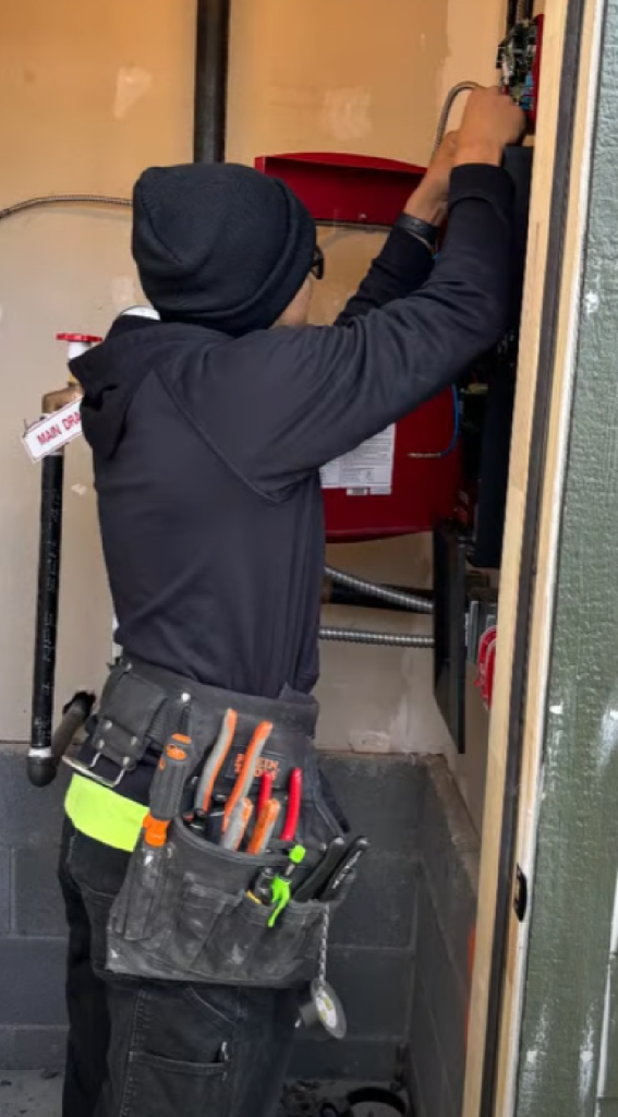 A technician wearing a tool belt works on a fire alarm control panel inside a utility area, adjusting wiring while standing near a red system cabinet and piping.