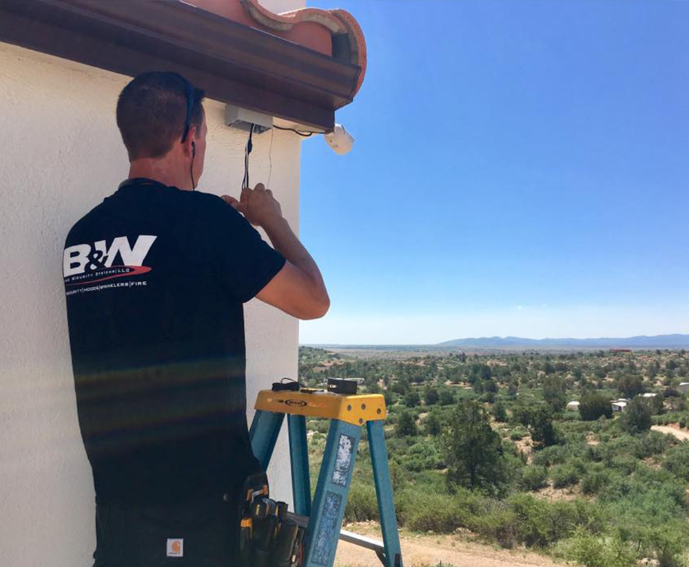 A technician wearing a B&W-branded shirt stands on a ladder installing or adjusting a security camera mounted under the roof eave of a building. The scene overlooks a wide, sunlit landscape with scattered trees, homes, and distant hills under a clear blue sky.