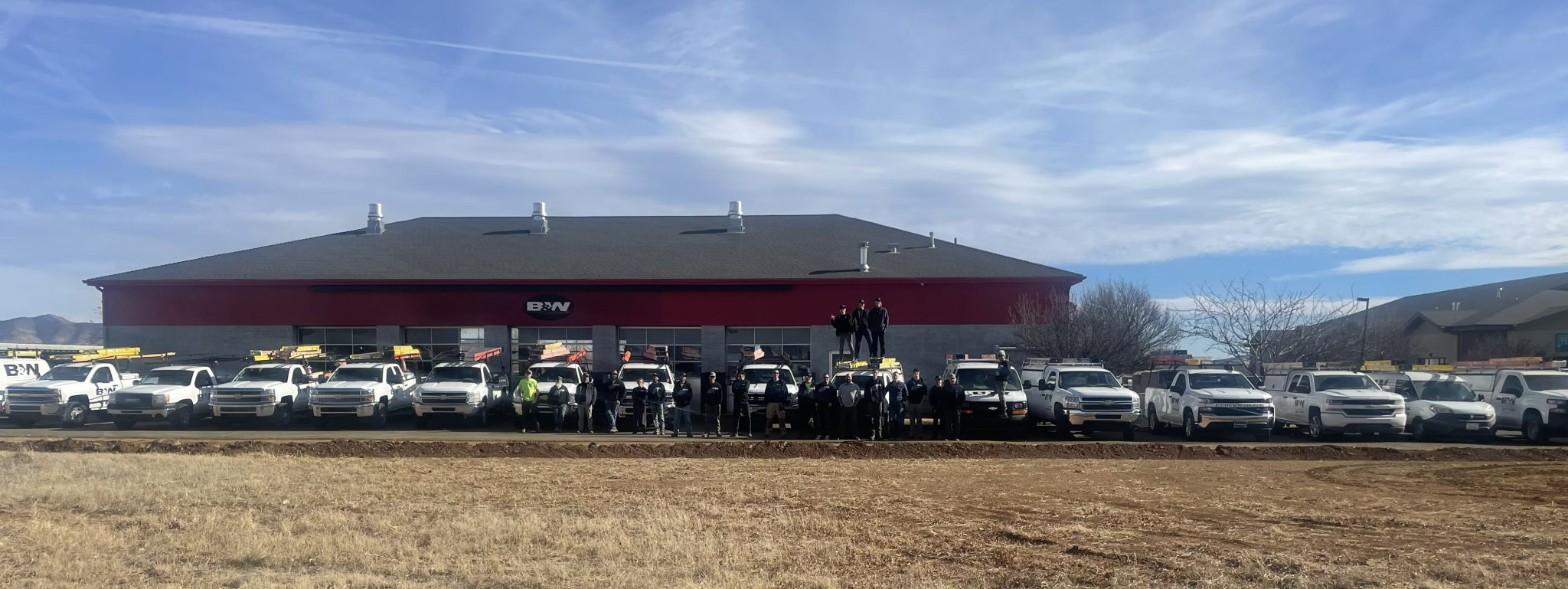 A wide outdoor group photo shows a team of workers standing in front of a large commercial building with a red upper facade and gray lower walls. A row of white service trucks, many equipped with ladders on top, is parked in front of the building. Several people stand on top of one of the trucks while others line up along the ground. The building features a central sign with a company logo, and the scene is set under a bright blue sky with scattered clouds, with dry grass and open land in the foreground.