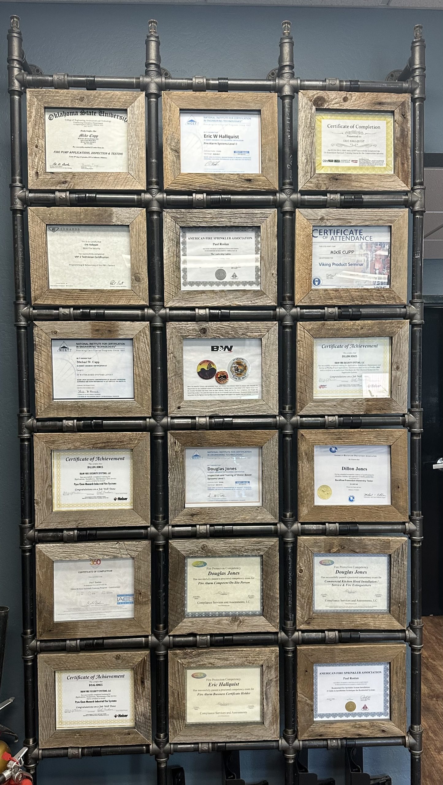 A wall display made from dark metal pipes holds a grid of rustic wooden frames, each containing a professional certificate. The certificates appear to be related to fire protection, alarm systems, and safety training, with various names and organizations visible. The frames are arranged in neat rows and columns, creating an industrial-style feature wall against a blue-gray painted surface.